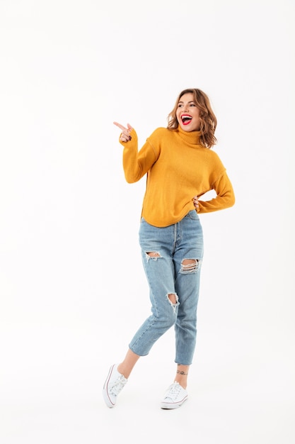 Full length Cheerful woman in sweater posing with arm on hip hile pointing and looking away over white wall