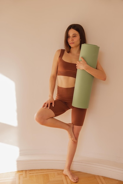 Foto gratuita giovane ragazza caucasica sicura di tutta la lunghezza che tiene un tappetino da yoga contro il muro bianco la sportiva dai capelli scuri indossa un top marrone e leggings in allenamento concetto di stile di vita attivo sano