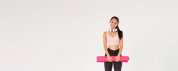 Full length of cute asian girl like fitness holding rubber mat for exercises or yoga classes standin