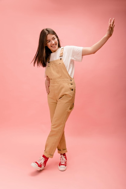 Free photo full length cute young caucasian brunette in casual clothes waving to side on pink background concept of friendliness