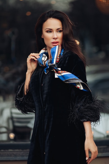 Free photo full length of a fashionable lady in black coat and leather boots wearing colorful handkerchief around her neck. she is carrying a cup of takeaway coffee against open car in the street.