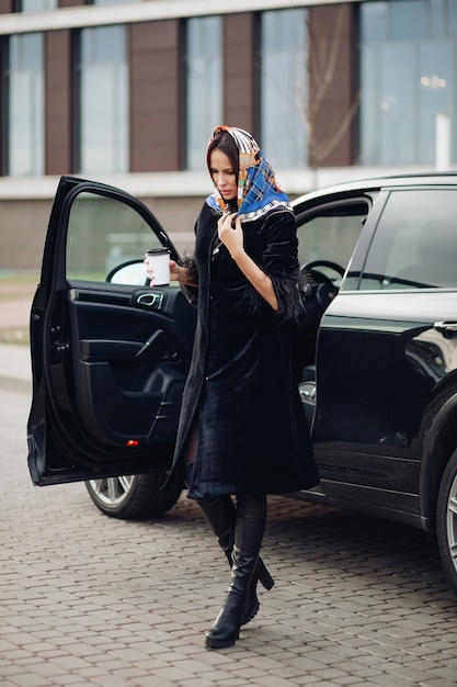 Full length of a fashionable lady in black coat and leather boots wearing colorful handkerchief around her neck. She is carrying a cup of takeaway coffee against open car in the street.