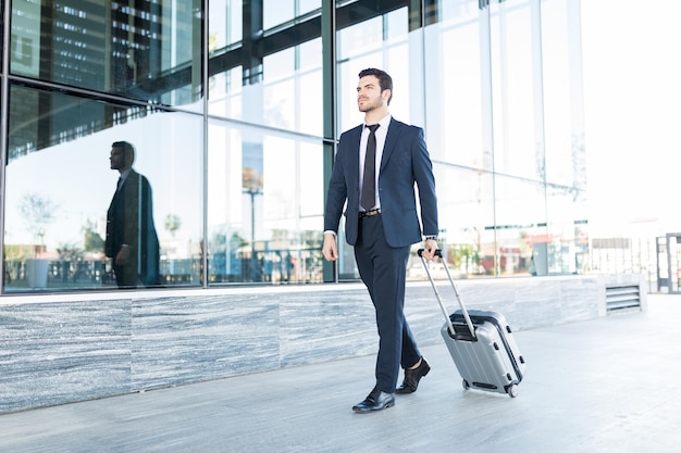 Full length of handsome young man in suit walking with luggage outside office building