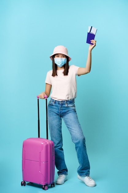 Full length of happy asian tourist girl travelling abroad posing with her suitcase in medical face m...