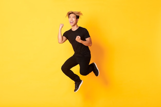 ジャンプして勝利を祝う黒い服を着た幸せな魅力的なアジアの男のフルレングスは、目標を達成し、黄色の壁に立って、勝利します。