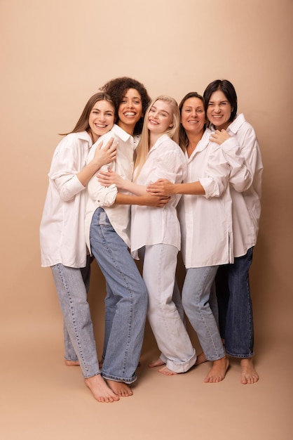 Signore intere diverse felici a figura intera in camicie bianche e jeans che sorridono alla telecamera su sfondo beige