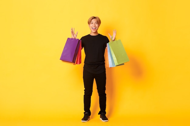 Foto gratuita integrale del ragazzo asiatico bello felice sullo shopping, tenendo le borse e sorridente, parete gialla.