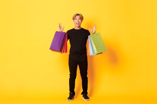В полный рост счастливый красивый азиатский парень по магазинам, держит сумки и улыбается, желтая стена.