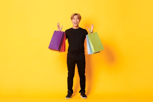 Full-length of happy handsome asian guy on shopping, holding bags and smiling, yellow wall.