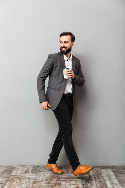 Full-length happy man with takeaway coffee smiling, and walking along gray