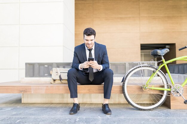 市内で自転車に乗って座っている間、携帯電話でテキストメッセージを入力するビジネス服装のヒスパニック系男性の全身