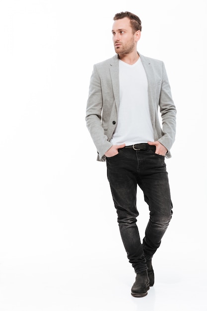 Full-length image of brunette businessman in jacket confidently posing with hands in pockets and looking aside, isolated over white wall