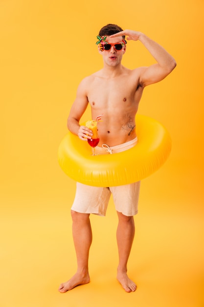 Full length image of Carefree naked man in shorts, unusual sunglasses and inflatable ring holding cocktail 