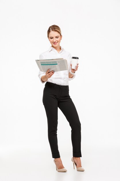 Full length image of Cheerful blonde business woman reading newspaper while holding cup of coffee over white wall