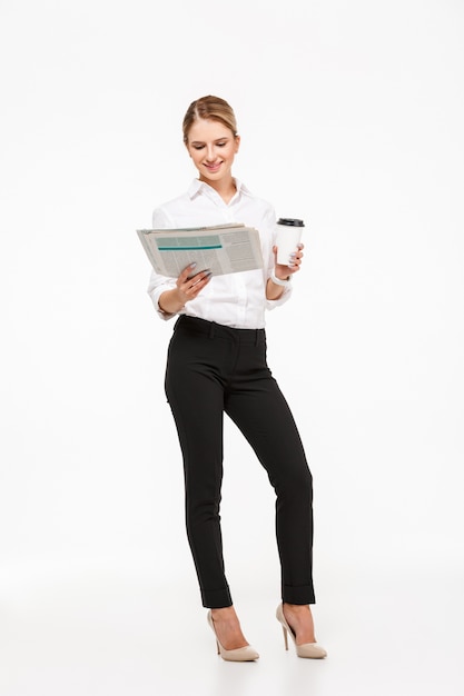 白い壁にコーヒーカップを押しながら新聞を読んで陽気な金髪ビジネス女性の完全な長さの画像
