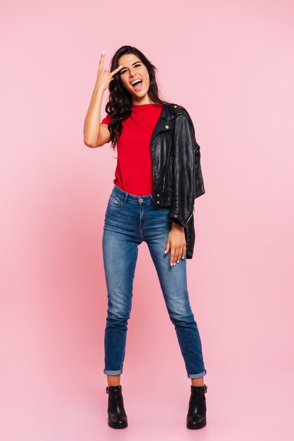 Full length image of cool screaming brunette woman showing rock gesture and looking at the camera over pink 