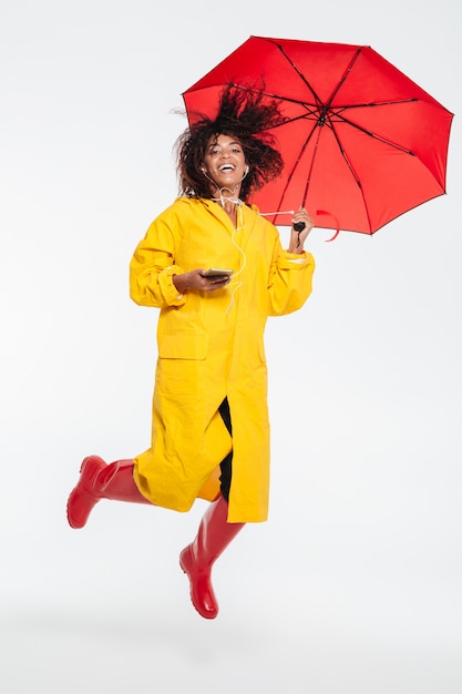 Изображение во всю длину счастливой африканской женщины в плаще, прячущемся под зонтиком и прыгающем во время прослушивания музыки белого цвета