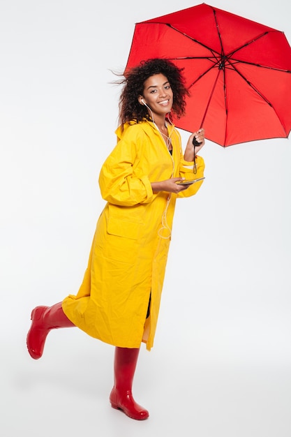 Полнометражное изображение счастливой африканской женщины в плаще прячется под зонтиком и слушать музыку на белом фоне