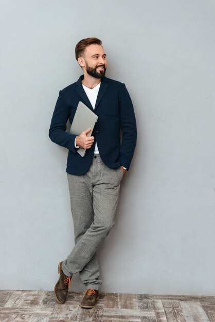 Full length image of happy bearded man in busines clothes holding laptop computer and looking awayy over gray 