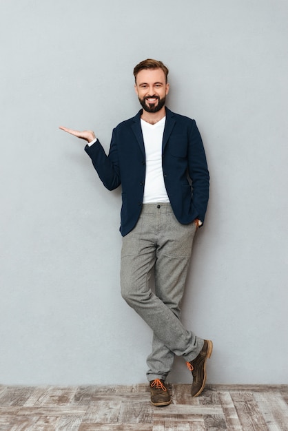 Full length image of happy bearded man in busines clothes with arm in pocket holding copyspace on the pound and looking at the camera over gray 