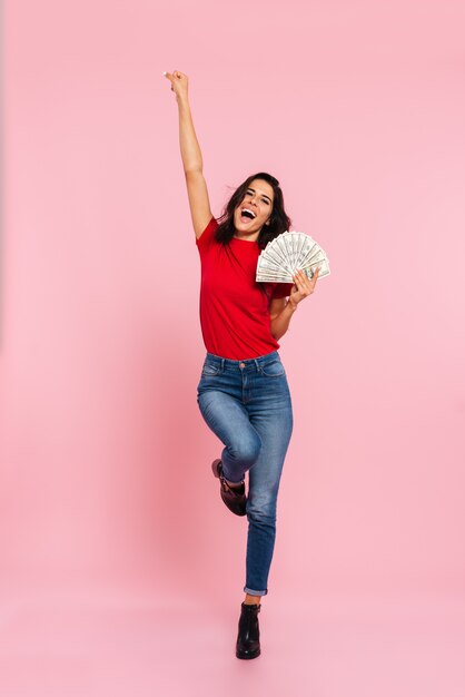 Full length image of happy brunette woman rejoice and holding money while looking at the camera over pink 
