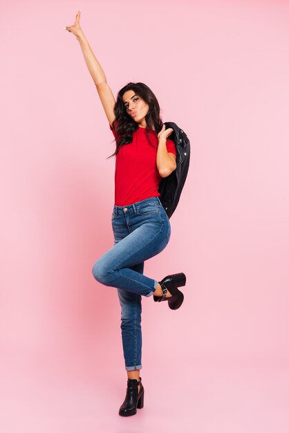 Full length image of pretty brunette woman posing in studio and showing peace sign while looking at the camera over pink 