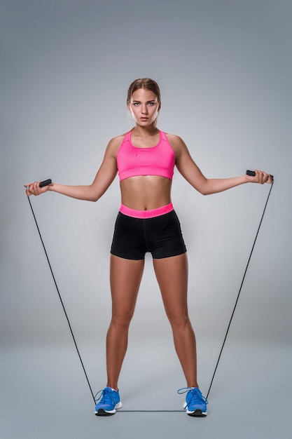 Full length image of a pretty fitness woman doing exercise with skipping rope over gray background. Young woman with beautiful slim healthy body posing in studio.