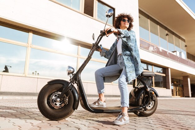 Полнометражное изображение милой усмехаясь курчавой женщины в солнечных очках сидя на современном мотоцикле outdoors и смотря прочь