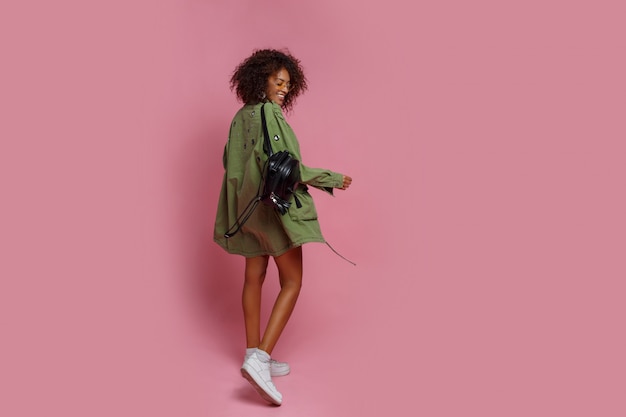 ピンクの背景のスタイリッシュな緑のジャケットに褐色肌の格好の良い女性の完全な長さの画像。ショッピングとファッションのコンセプトです。