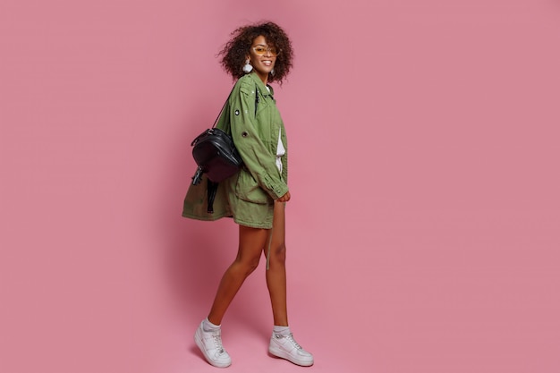 Full length image of shapely woman with dark skin in stylish green jacket on pink background. Shopping and fashion concept.