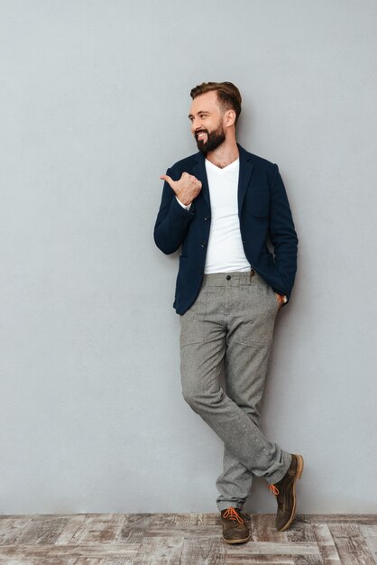 Full length image of smiling bearded man in busines clothes holding arm in pocket while pointing and looking away over gray 