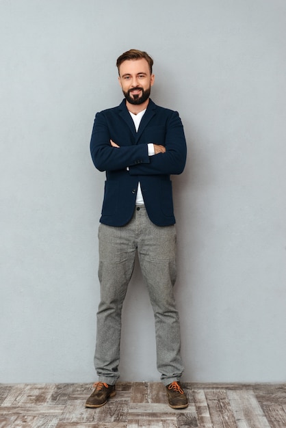 Full length image of smiling bearded man in busines clothes posing with crossed arms and looking at the camera over gray 
