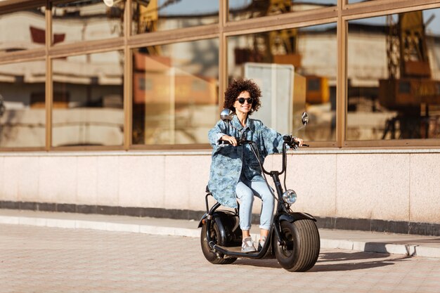 サングラスで笑顔の巻き毛の女性の完全な長さの画像は屋外の現代のバイクに乗る