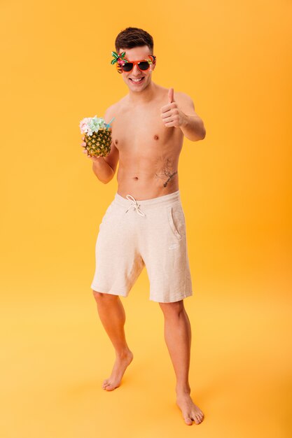 Full length image of Smiling naked man in shorts and unusual sunglasses holding cocktail while showing thumb up 