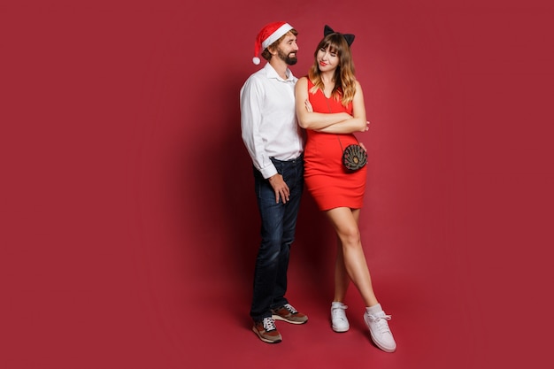Full length image of stylish couple in love in new year masquerade hats standing on red.