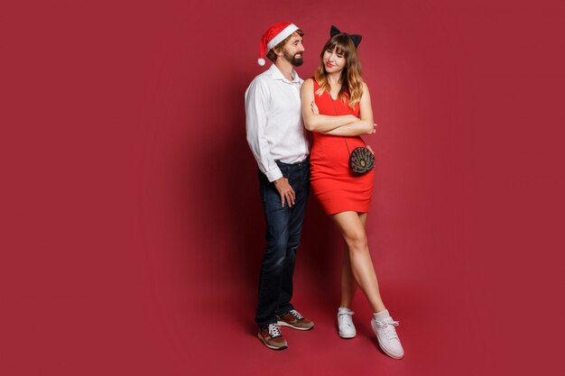 Полнометражное изображение стильной пары в влюбленности в шлемах маскарада Нового Года стоя на красном цвете.