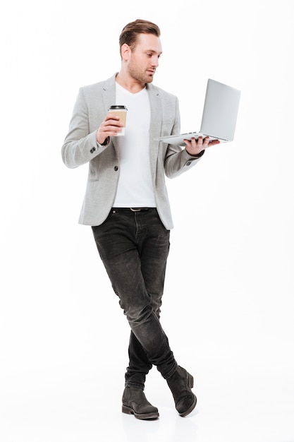 Full-length image of young entrepreneur in jacket standing with silver laptop and takeaway coffee in hands, isolated over white wall