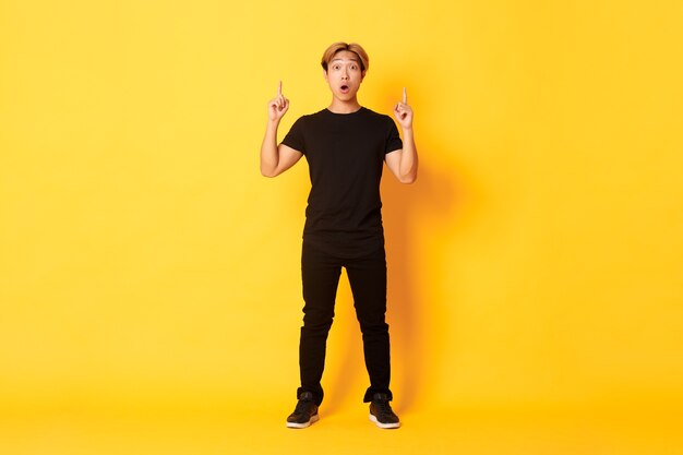 Full-length of impressed asian guy in black clothes, gasping amazed and pointing fingers up, yellow wall