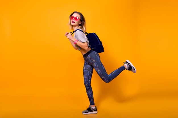 Full-length indoor portrait of girl wearing leggings, running on yellow background. Pretty slim female model in sneakers with backpack fooling around in studio.