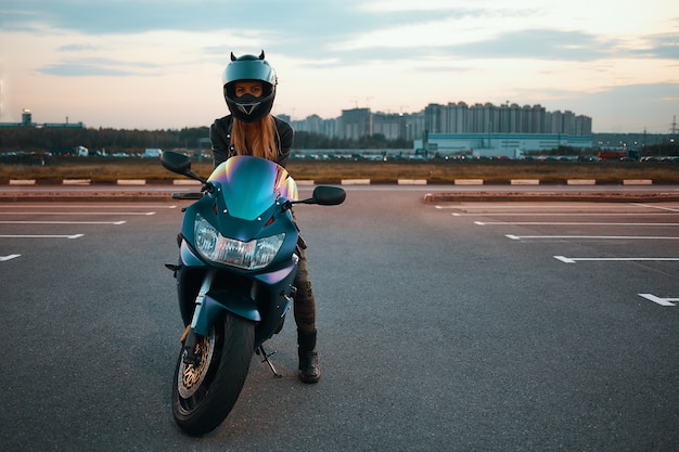 Full length isolated picture of fashionable active young female with blonde hair wearing safety helmet posing against multi storied buildings, sitting on motorbike with one foot on pavement