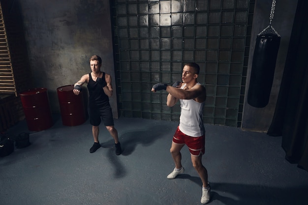 Full length isolated portrait of two handsome Caucasian guys exercising indoors together wearing stylish sports clothes and boxing bandages, reaching out hands while mastering punches in modern gym