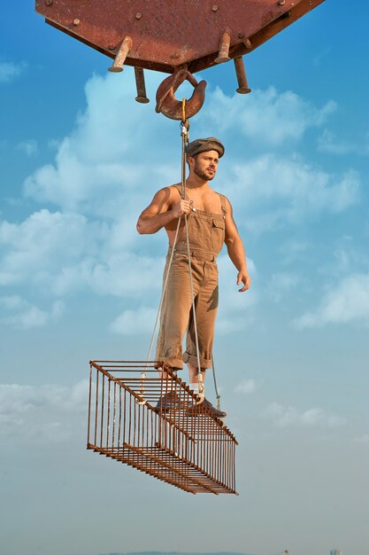 Full length of man holding and standing on iron construction on high at city. Builder wearing hat and work wear looking away and posing. Crane holding construction. Blue sky with clouds on background.