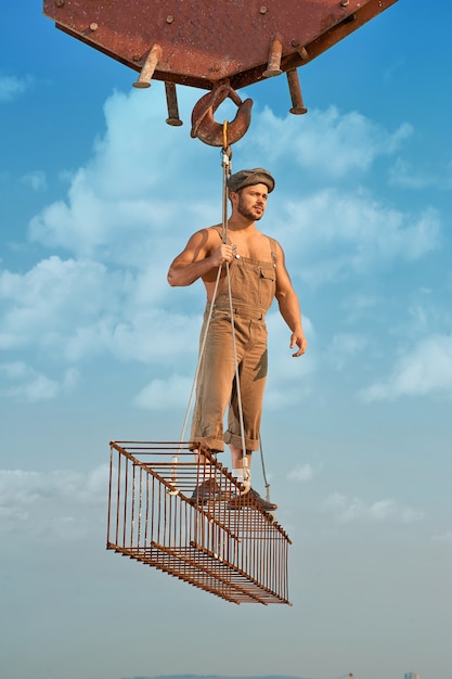 Full length of man holding and standing on iron construction on high at city. Builder wearing hat and work wear looking away and posing. Crane holding construction. Blue sky with clouds on background.