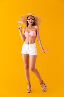黄色の壁の上に孤立して立っているビキニを着て、踊りながらカクテルを保持している魅力的な陽気な少女の全長