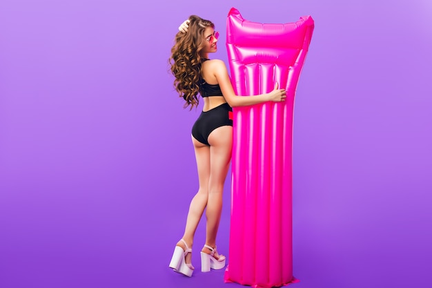 Full-length photo of attractive girl with long curly hair in pink sunglasses on purple background in studio. She wears swimsuit and holds pink air mattress near. View from back.