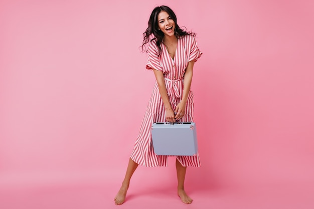 Full-length photo of cheerful lady dancing with suitcase in her hands. Tanned brunette with curls in midi length outfit posing barefoot.