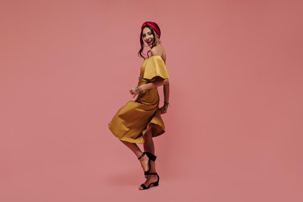 Full length photo of cheerful woman in midi yellow dress and black stylish heels dancing and looking into camera on pink backdrop