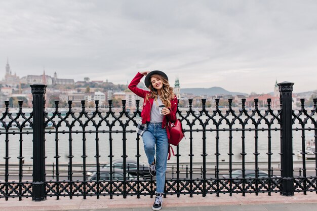 Full-length photo of confident woman in hat standing on one leg at bridge