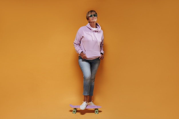 Full length photo of cool woman with short hair in sunglasses, wide hoodie and skinny jeans smiling and posing with pink skateboard