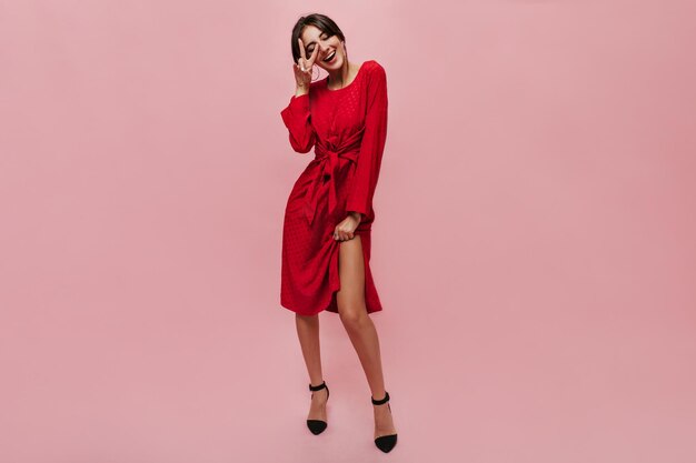 Full length photo of fashionable cool lady with round big earrings in midi red dress and black heels showing peace sign and smiling